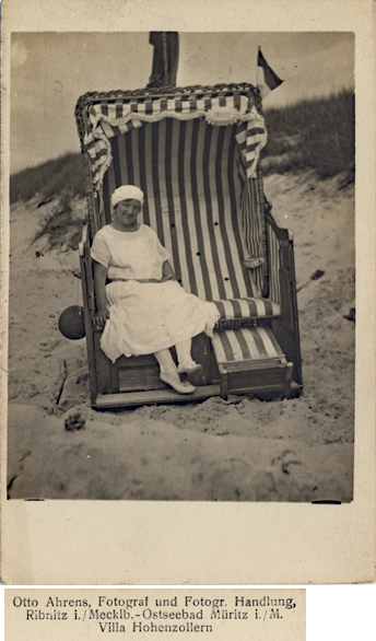 Handschriftlich auf 1925 datiert. Fotopostkarte. Rechts neben dem Kopf der Dame ist auf dem gestreiften Stoff der Name des Herstellers des Strandkorbes aufgedruckt: Johann Falck aus Rostock. 